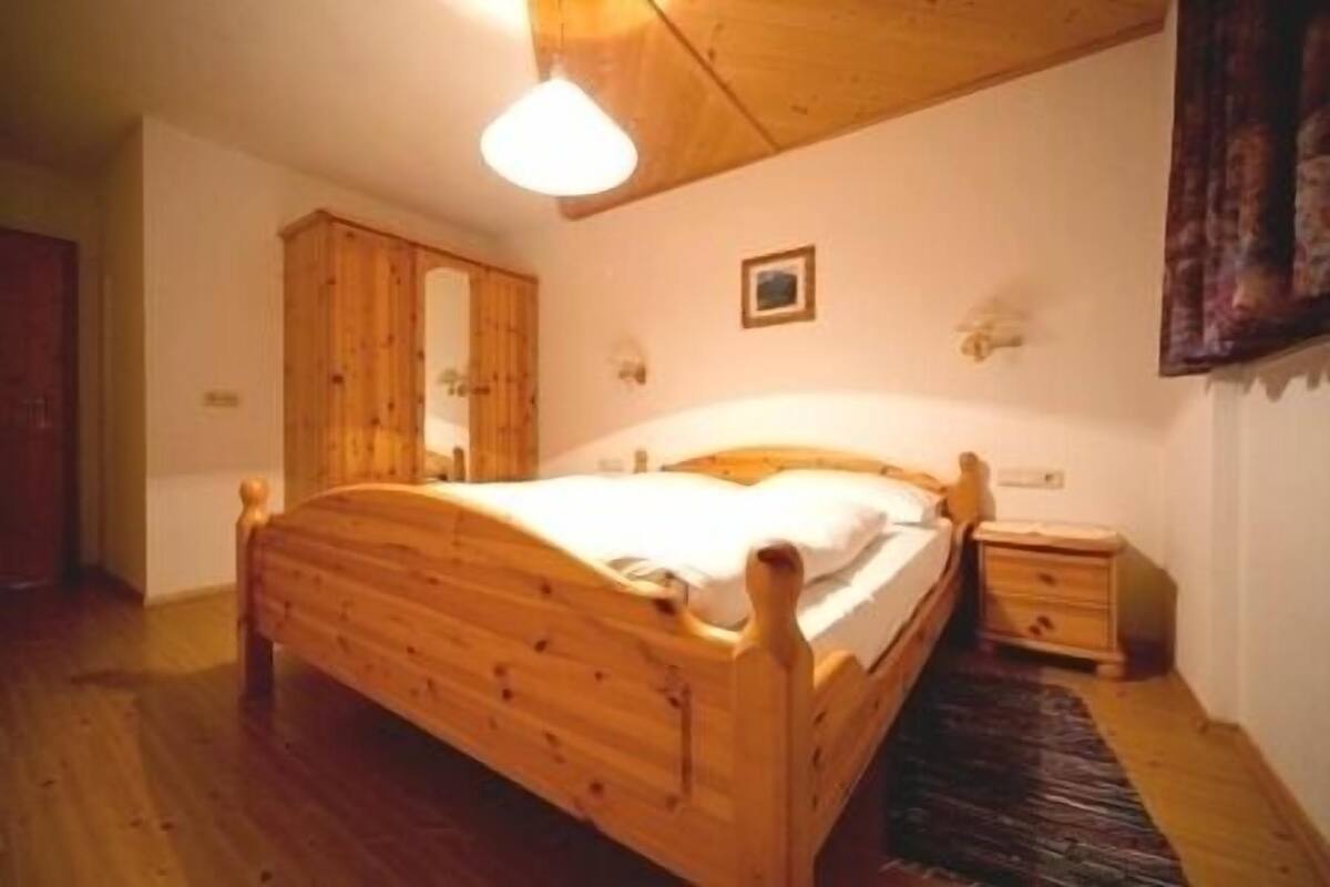 Una cama de madera en una habitación con techo y paredes de madera, una mesita de noche pequeña y una lámpara fijada en la pared.