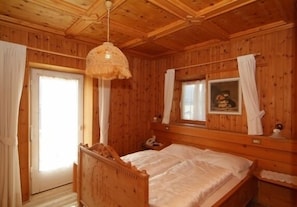 A wooden cabin room with a bed, a hanging light fixture, a window with curtains, and a painting on the wall.