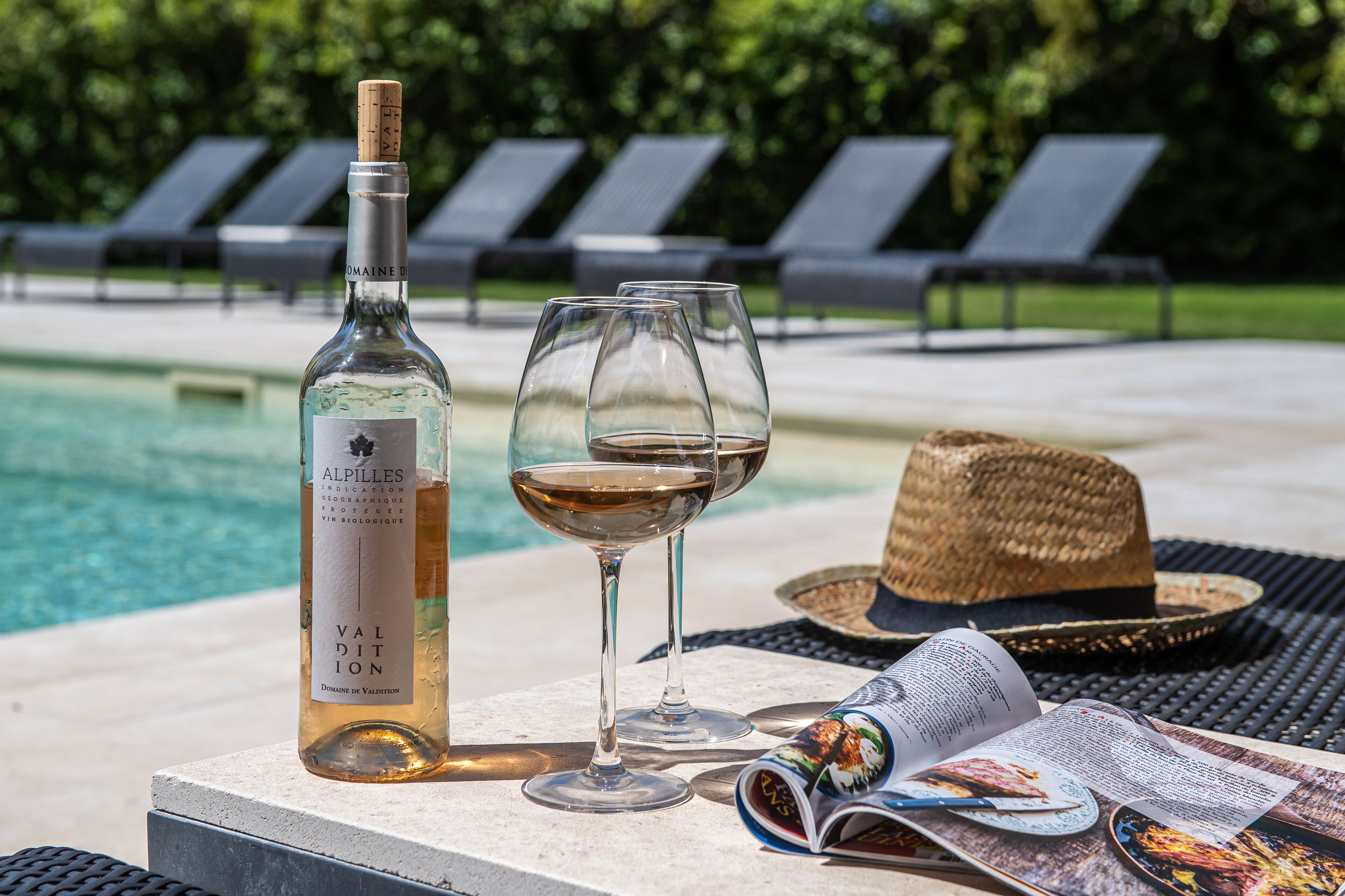 Une bouteille de vin, un verre à vin, un chapeau de paille et un magazine sur une table près d’une piscine.