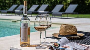 Une bouteille de vin, un verre à vin, un chapeau de paille et un magazine sur une table près d’une piscine.