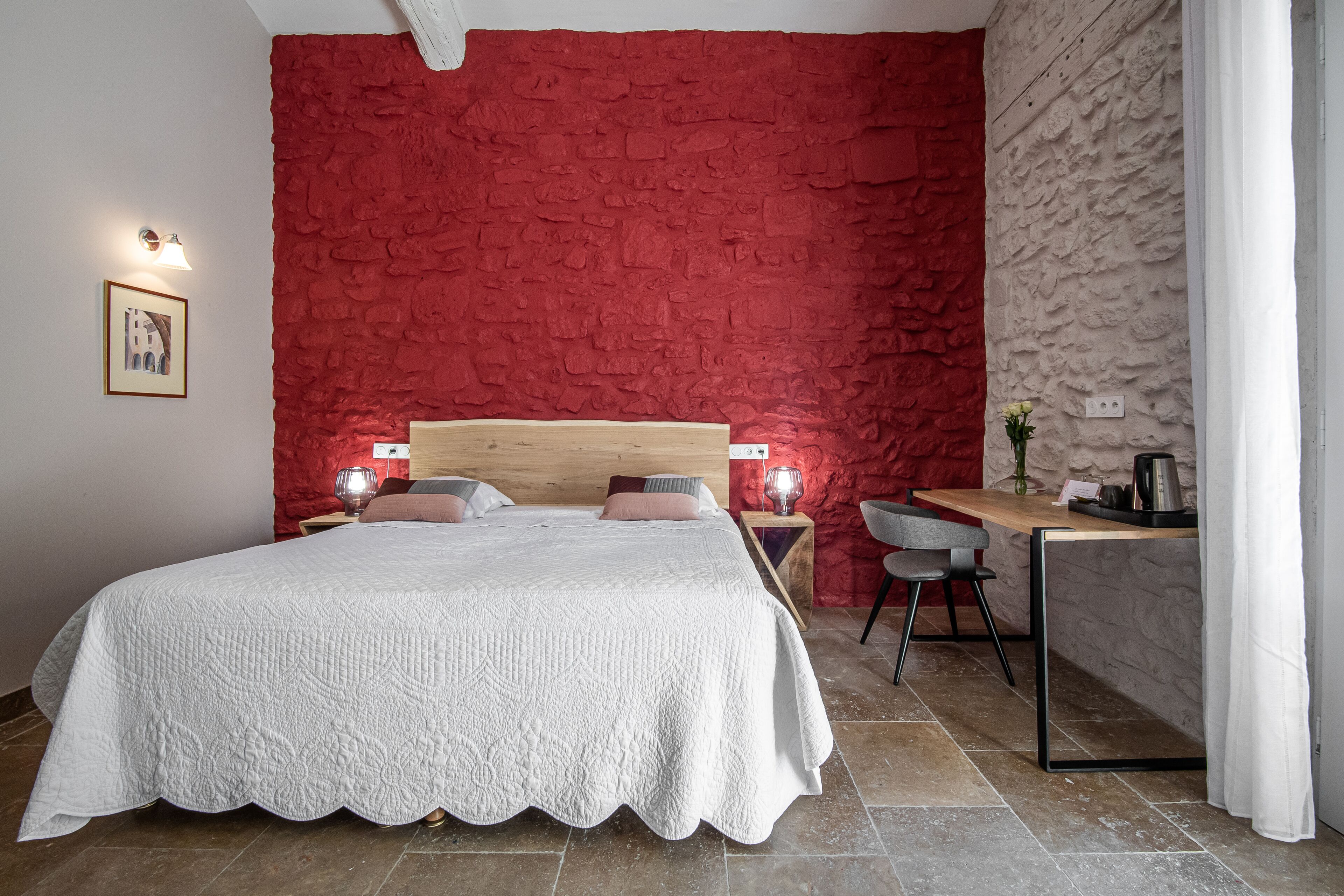 A bedroom with a red textured wall, a wooden headboard, a white quilted bedspread, two bedside lamps, a small table with a chair, and a window with white curtains.