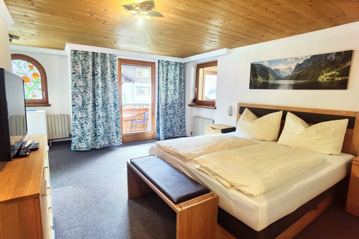 A bedroom with a wooden ceiling, a large bed with white bedding, a bench, a desk with a computer, and a window with curtains.