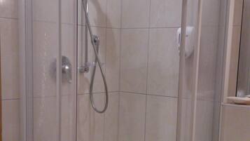 Ducha en esquina con mampara de vidrio, una toalla blanca en el suelo y un lavamanos con espejo.
