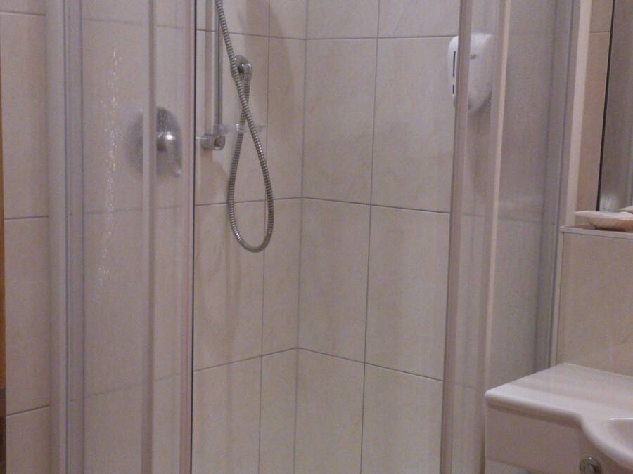 Ducha en esquina con mampara de vidrio, una toalla blanca en el suelo y un lavamanos con espejo.