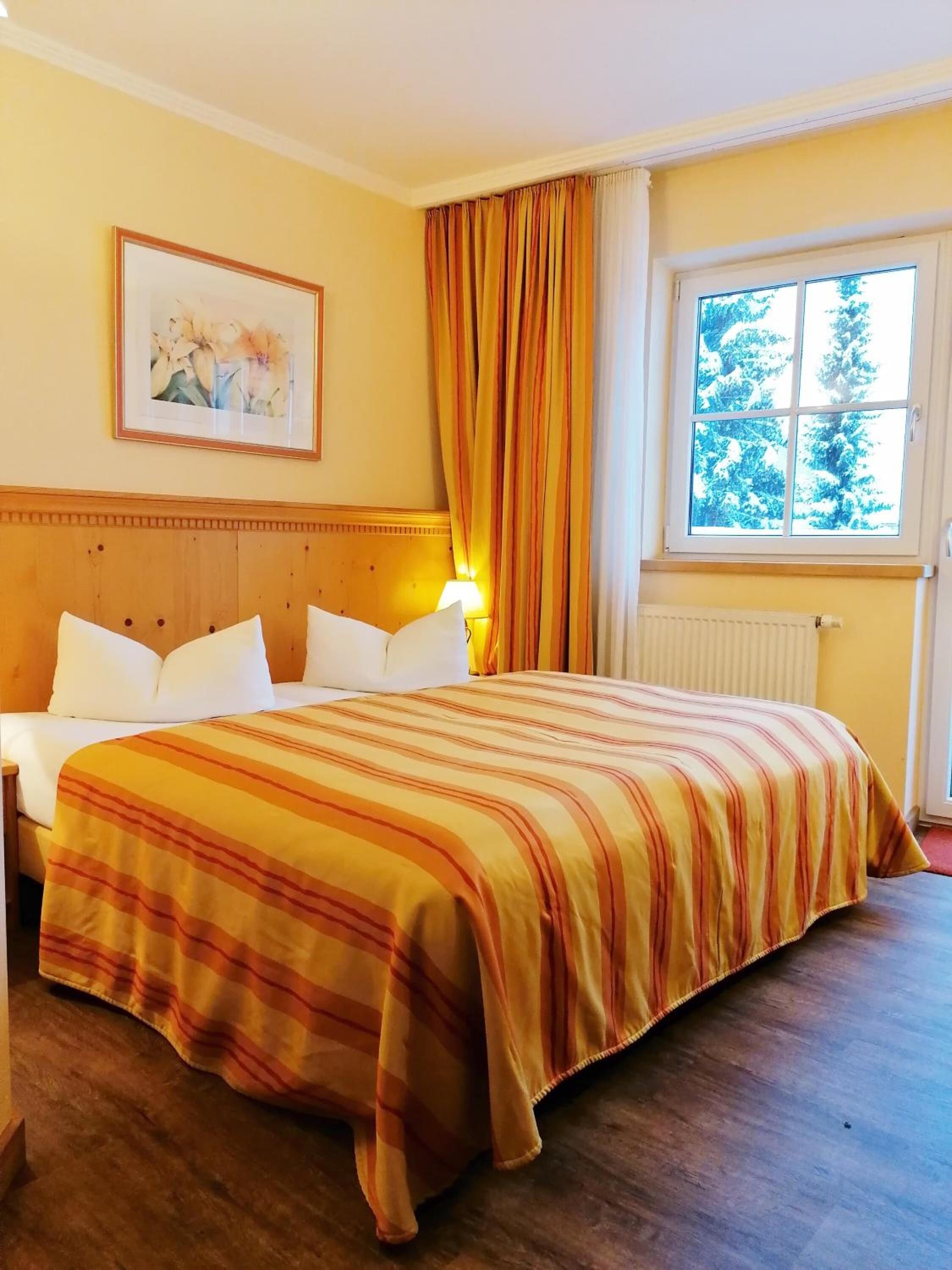 A hotel room with a bed, a headboard, a window with curtains, a radiator, and a framed picture on the wall.