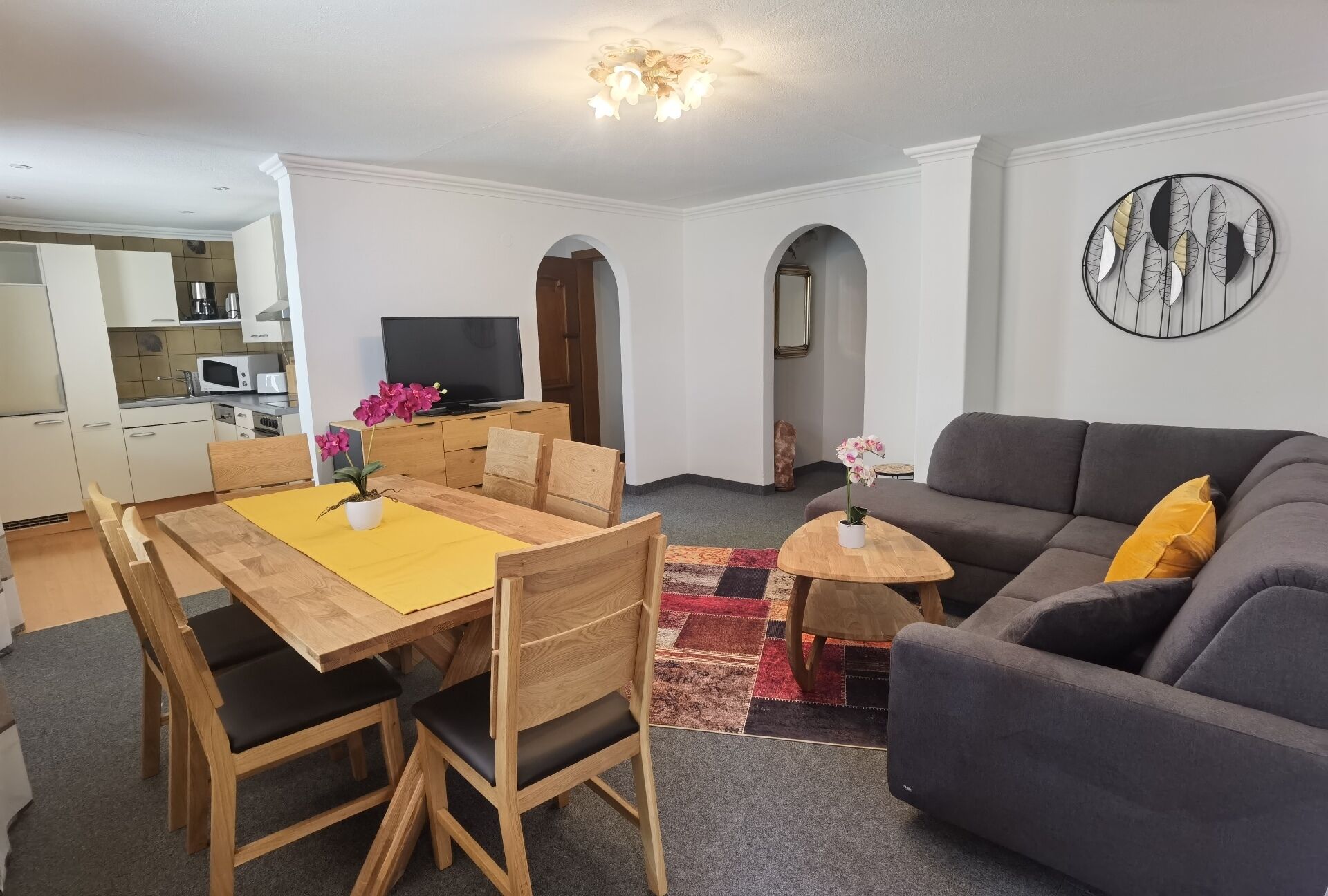 Un salon moderne avec un canapé gris, une table à manger en bois et des chaises. On y trouve également un coin cuisine avec des meubles blancs et un téléviseur à écran plat.