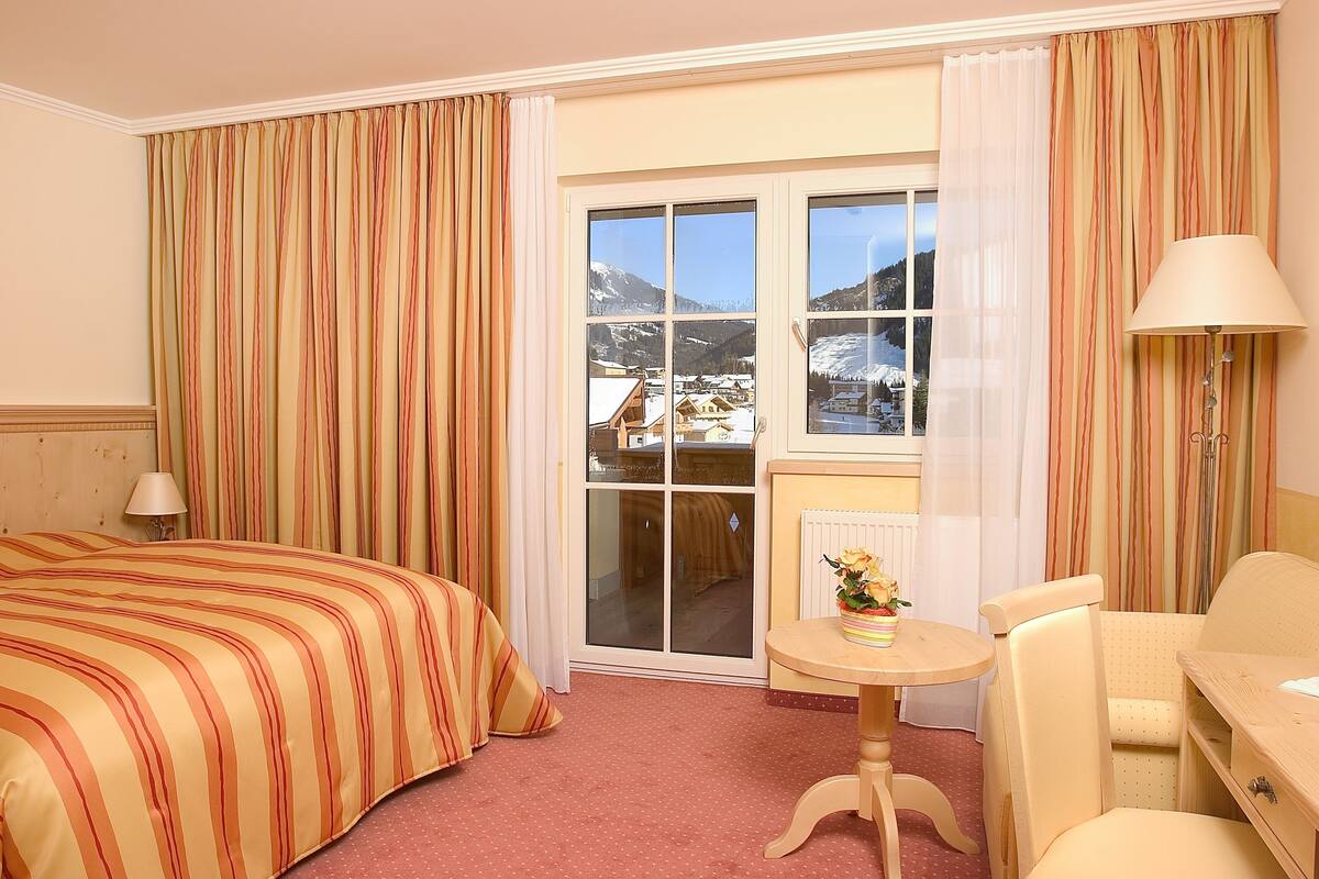 A hotel room with a bed, a small table with a lamp, a chair, and a view of snowy mountains through a window.