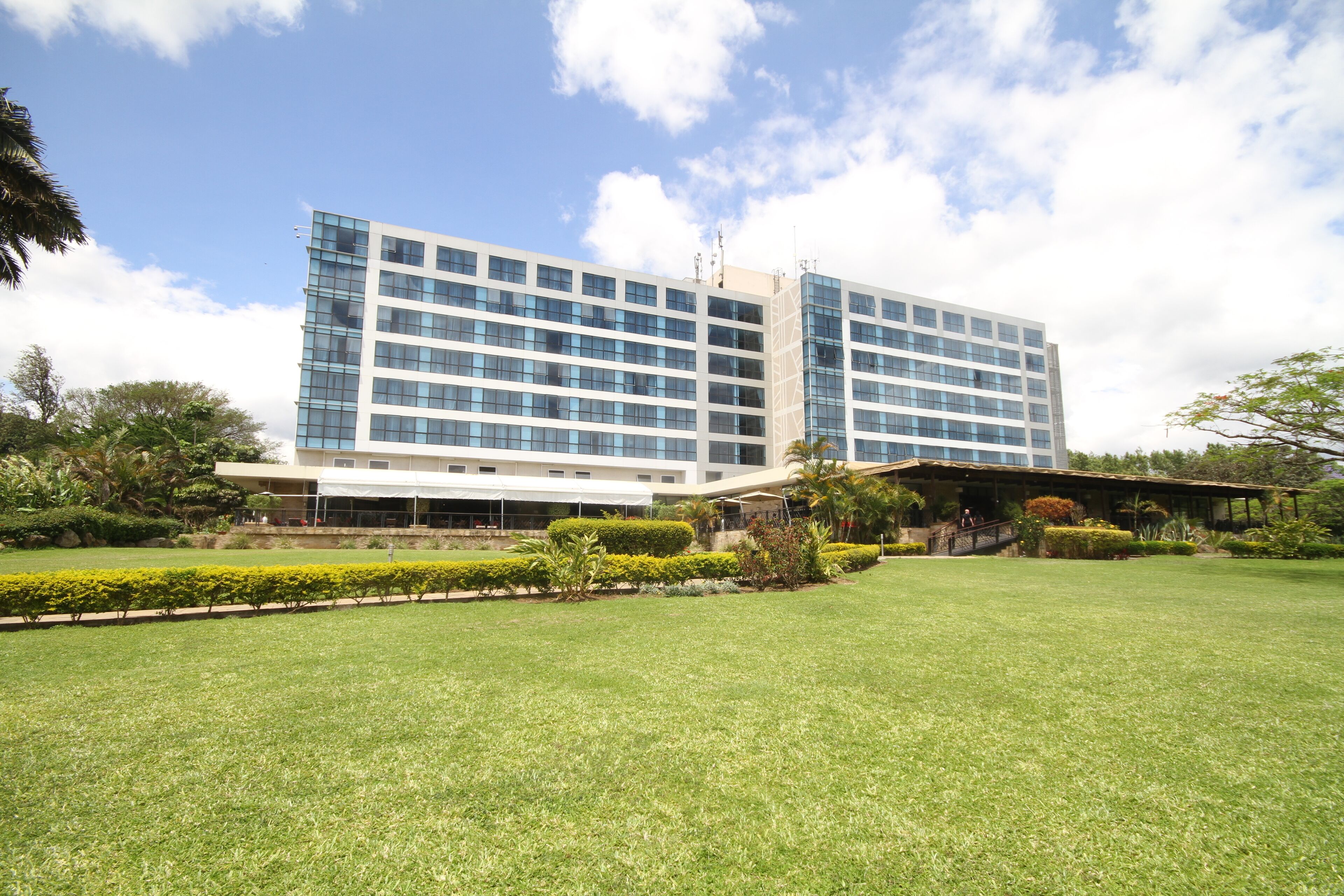 A modern hotel with a large glass facade, a well-maintained lawn, and a covered outdoor area with seating.