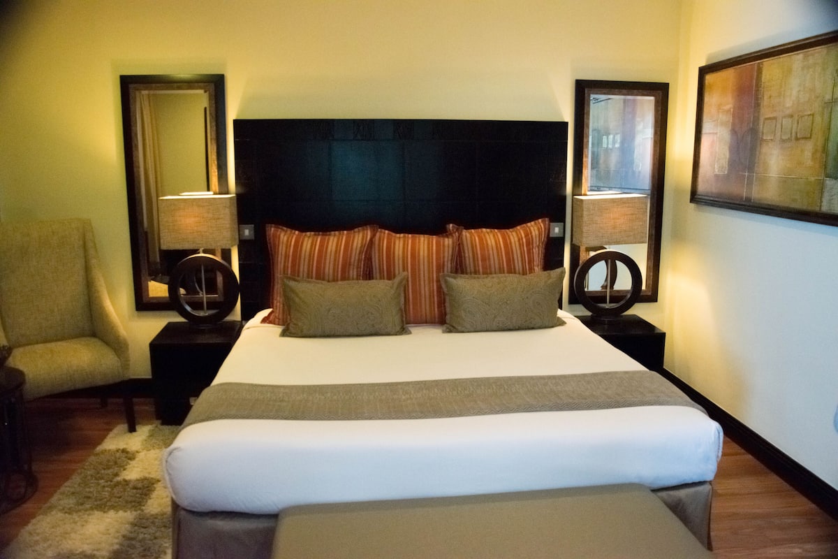 Una habitación de hotel con una cama grande, dos mesitas de noche con lámparas, una silla y un cuadro en la pared.