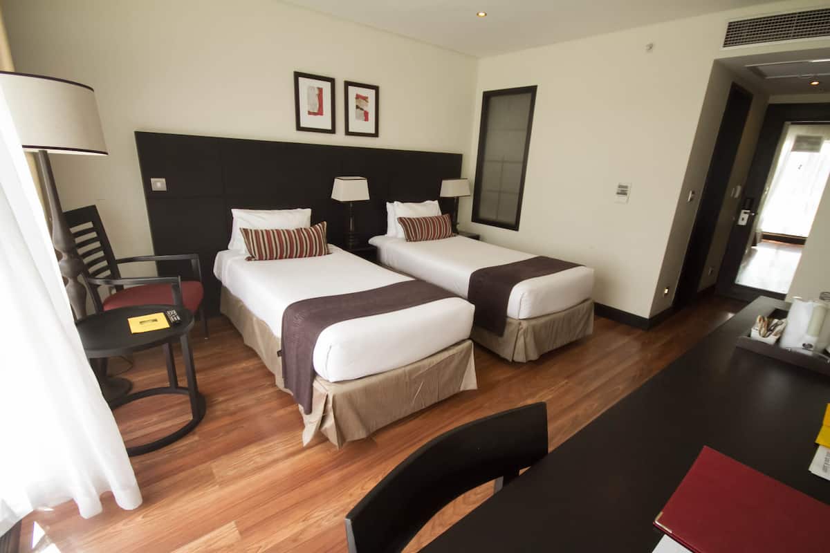 Una habitación de hotel con dos camas individuales, un escritorio con silla, una lámpara y una ventana.