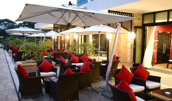 An outdoor seating area with wicker furniture and red cushions, surrounded by umbrellas and potted plants. - Mount Meru Hotel (Arusha)