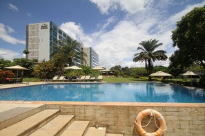 A swimming pool with lounge chairs and umbrellas, surrounded by palm trees and a building in the background.