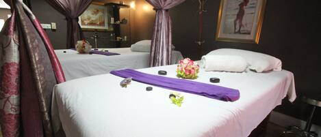 A hotel room with two beds, each with white linens and purple accents. There are decorative pillows, a vase with flowers, and a purple towel on one bed. The room has a dark wall with a framed picture, and a coat stand is visible.