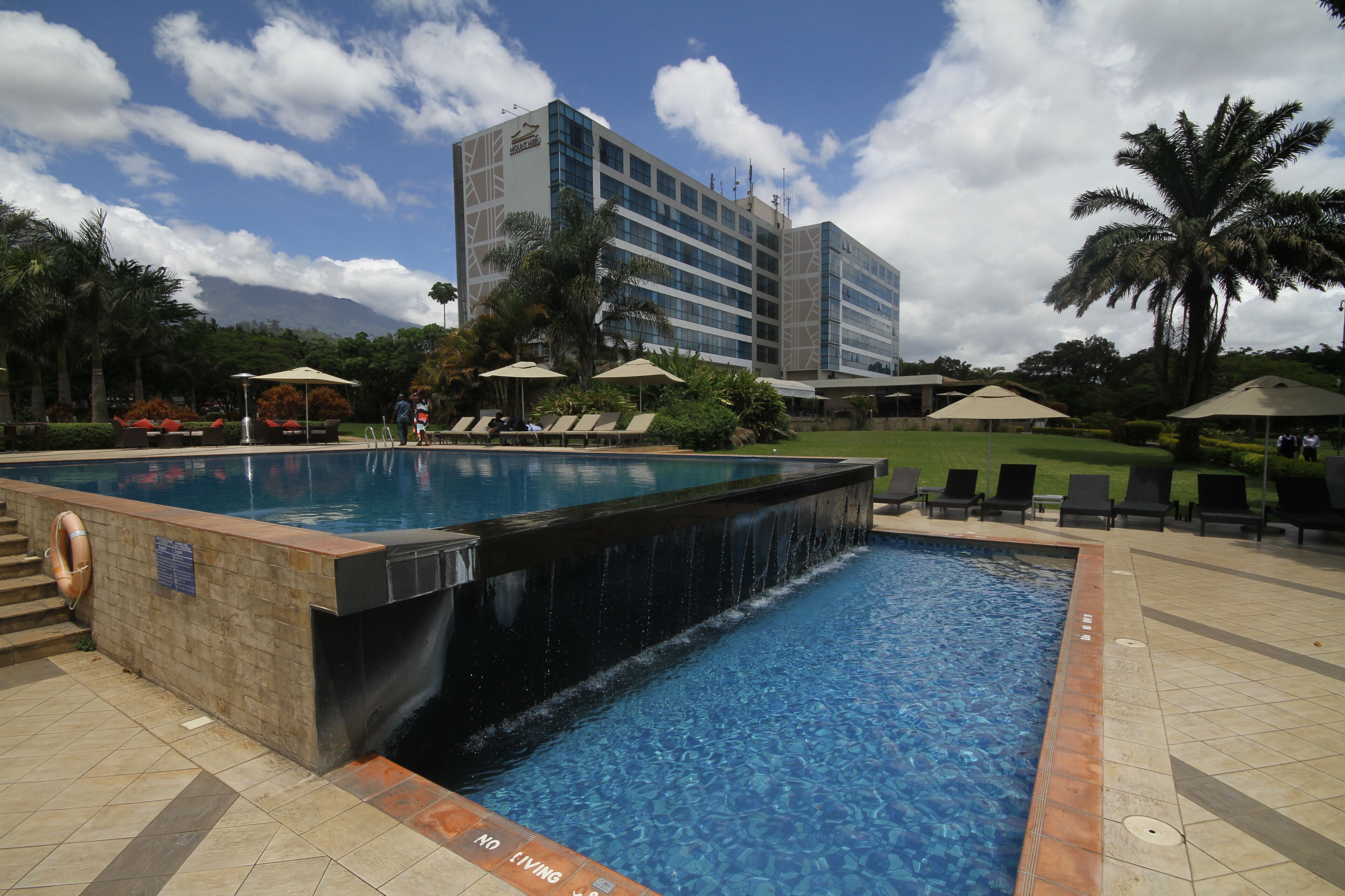 Una piscina con cascada, rodeada de sillones y sombrillas, con un edificio de hotel al fondo.