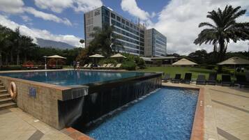 Una piscina con cascada, rodeada de sillones y sombrillas, con un edificio de hotel al fondo.