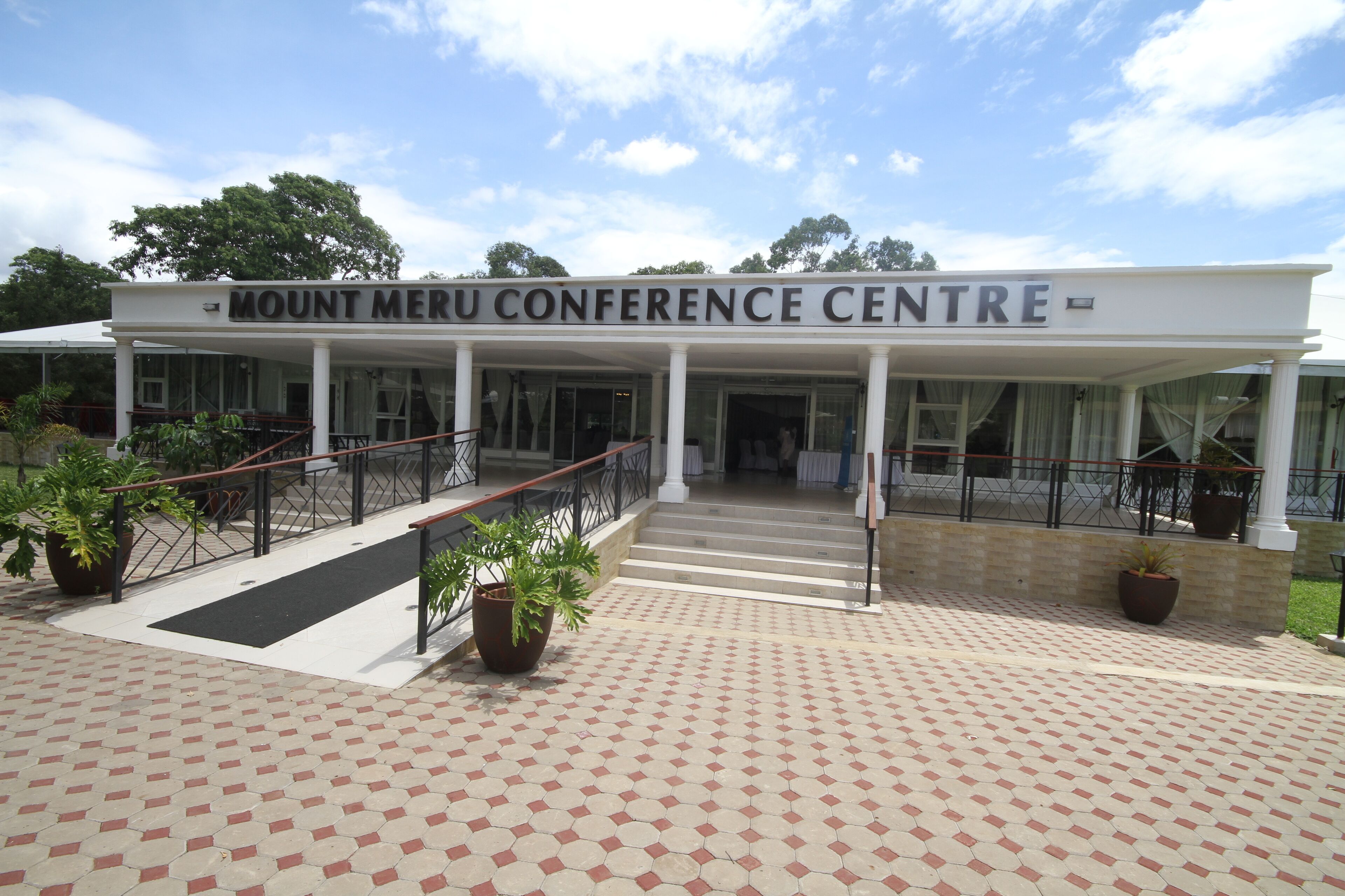 El centro de conferencias Monte Meru, con una entrada techada, un camino de baldosas y plantas en macetas.