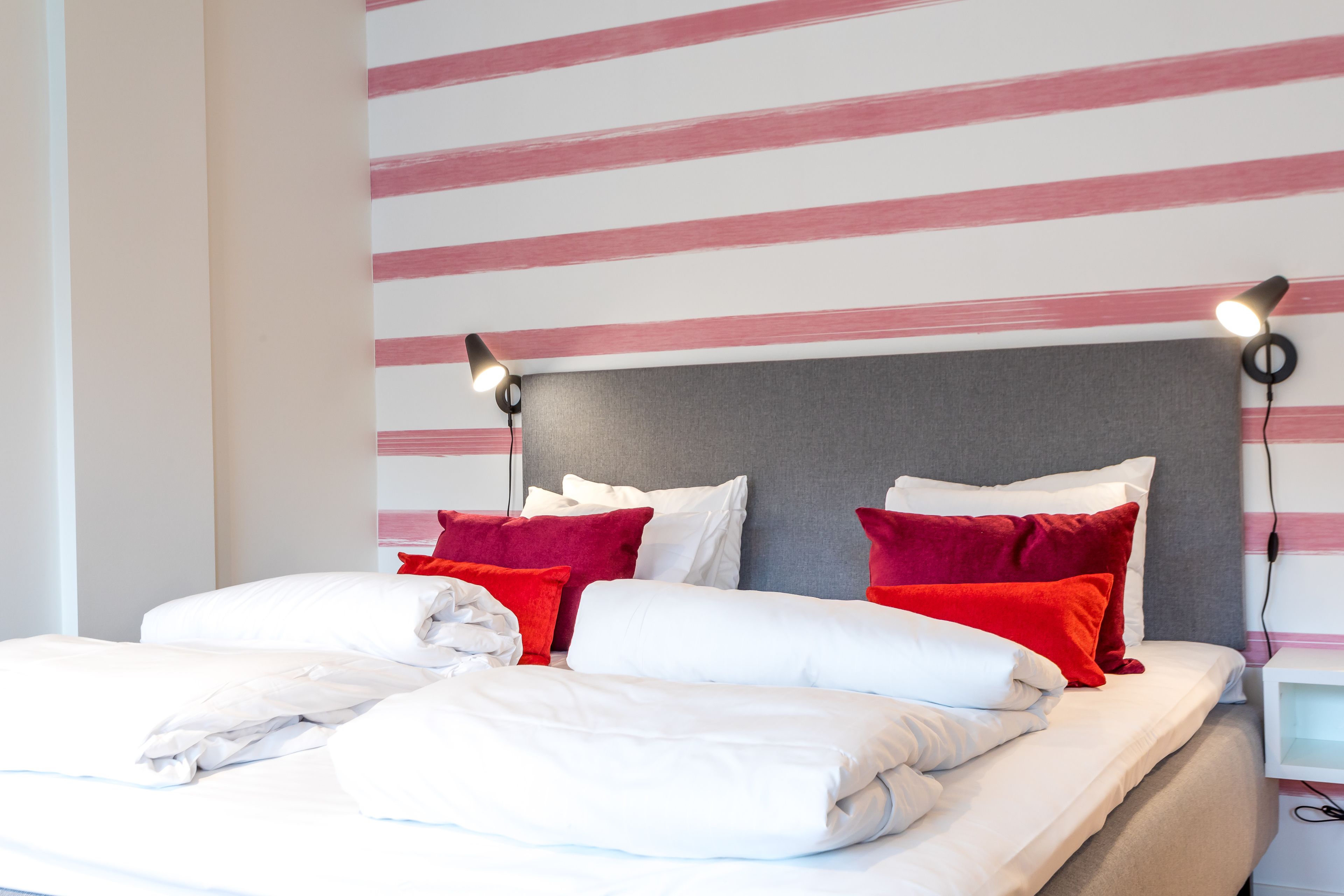 A neatly made bed with white and red pillows, a grey headboard, and two bedside lamps.