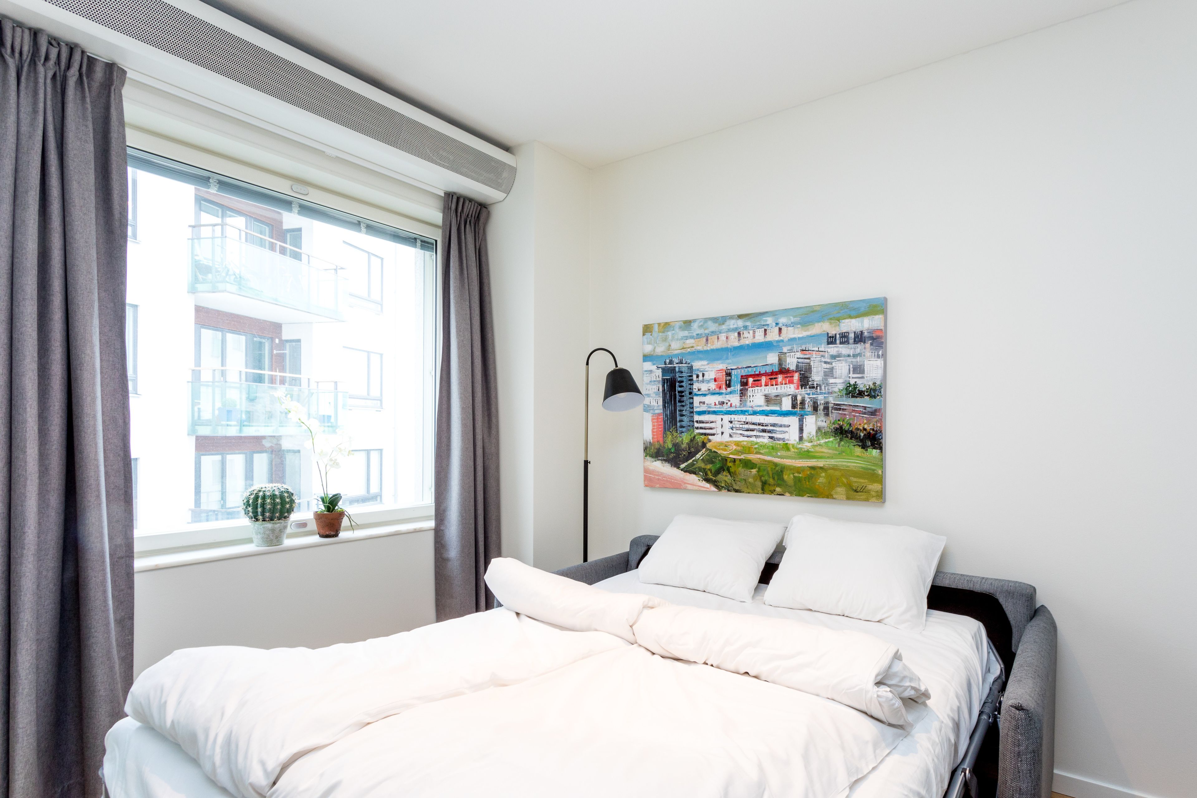 A neatly made bed with white bedding, a window with grey curtains, a small cactus and potted plant on the windowsill, and a colorful cityscape painting on the wall.