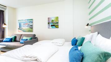 A modern hotel room with a grey sofa, a bed with white and blue bedding, and two wall-mounted paintings.
