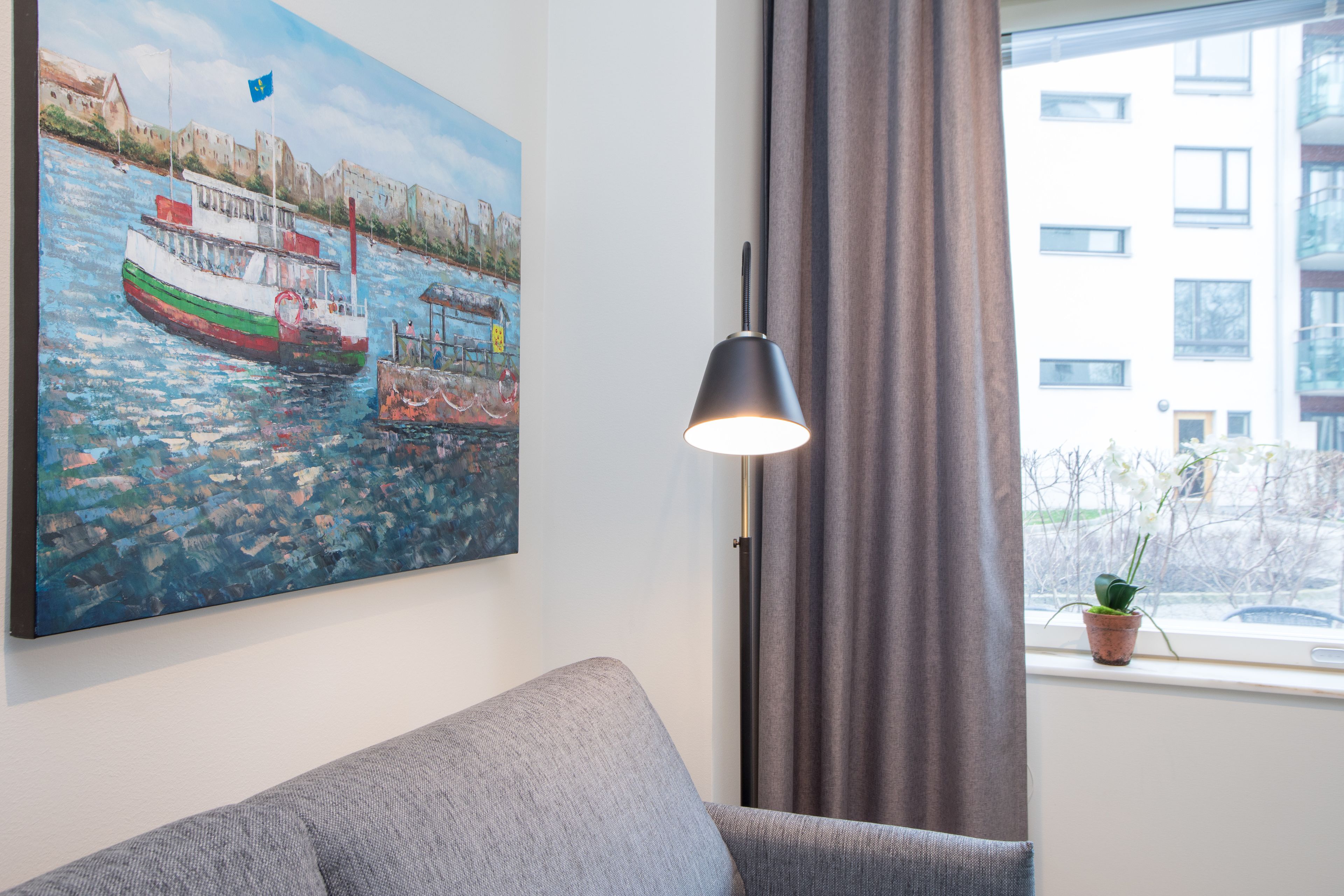 Una habitación con un cuadro de un barco en la pared, un sofá gris, una lámpara de pie y una ventana con cortina.