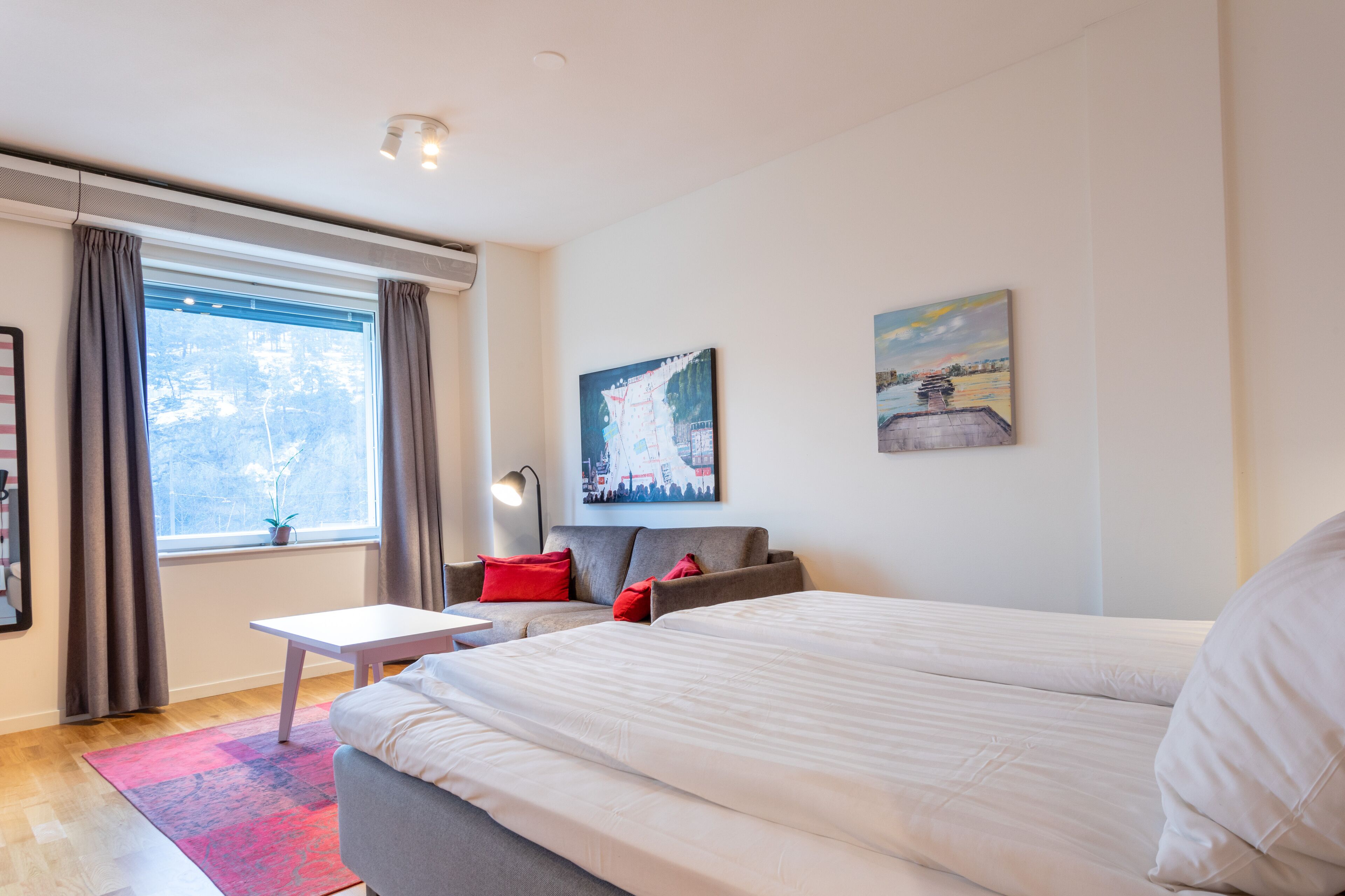 Ein modernes Hotelzimmer mit einem großen Bett, einem grauen Sofa, einem kleinen Tisch und einem Fenster mit Vorhängen.