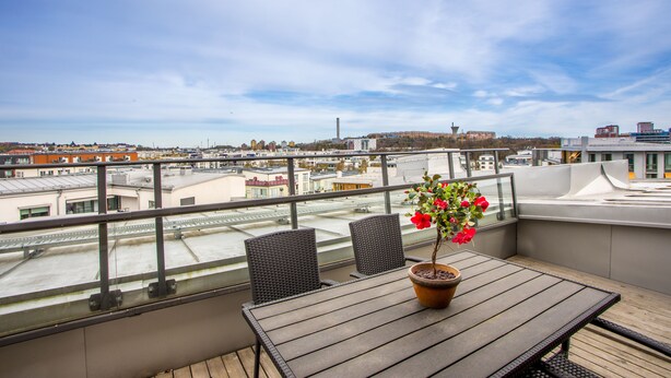 A balcony with a table and chairs, a potted plant with red flowers, and a city view. - ApartDirect Hammarby Sjöstad (Stockholm)