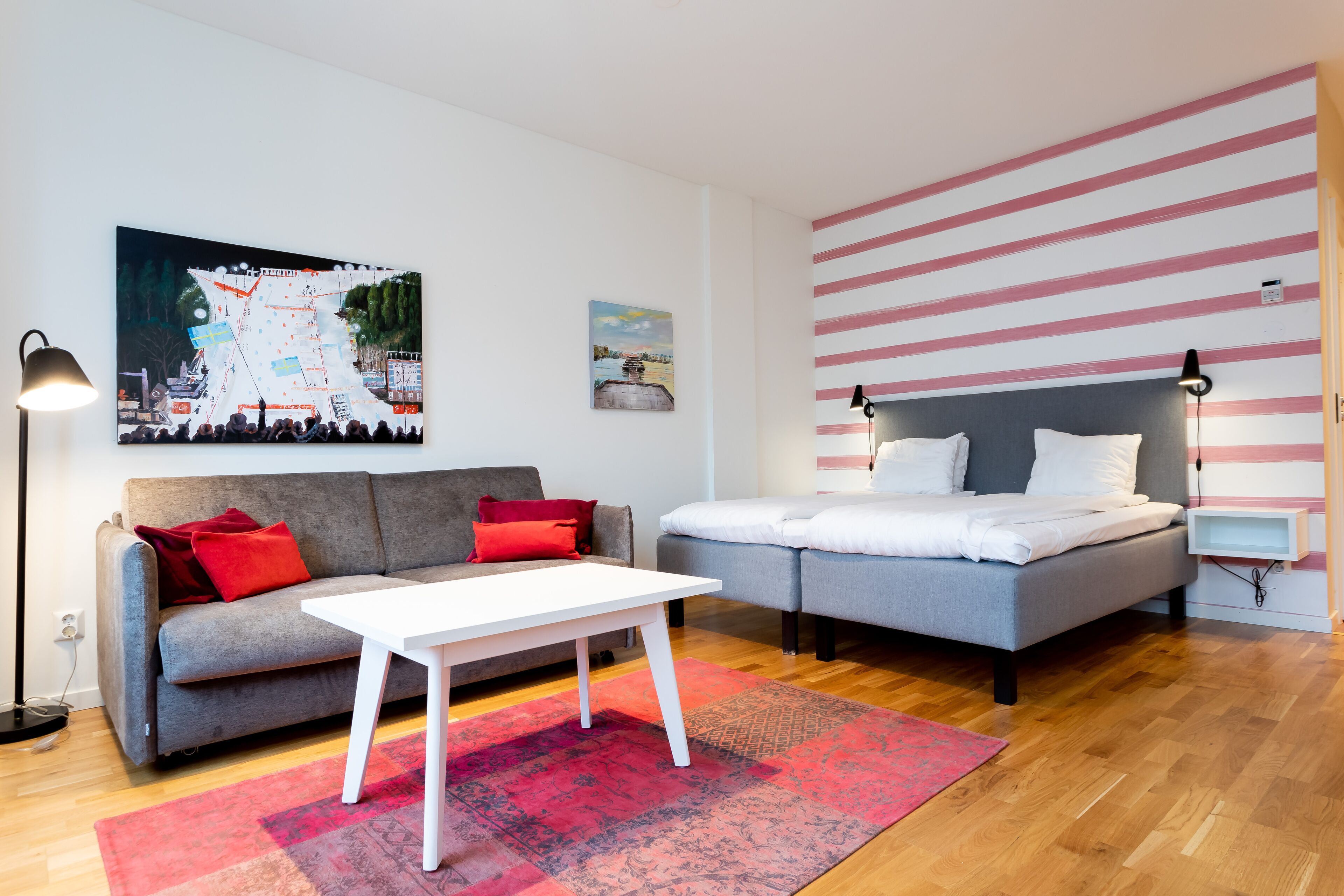 A modern living room with a grey sofa, a white coffee table, and a grey bed with white bedding. There is a red and pink patterned rug on the floor.