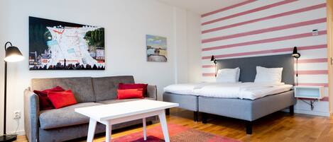 A modern living room with a grey sofa, a white coffee table, and a grey bed with white bedding. There is a red and pink patterned rug on the floor.
