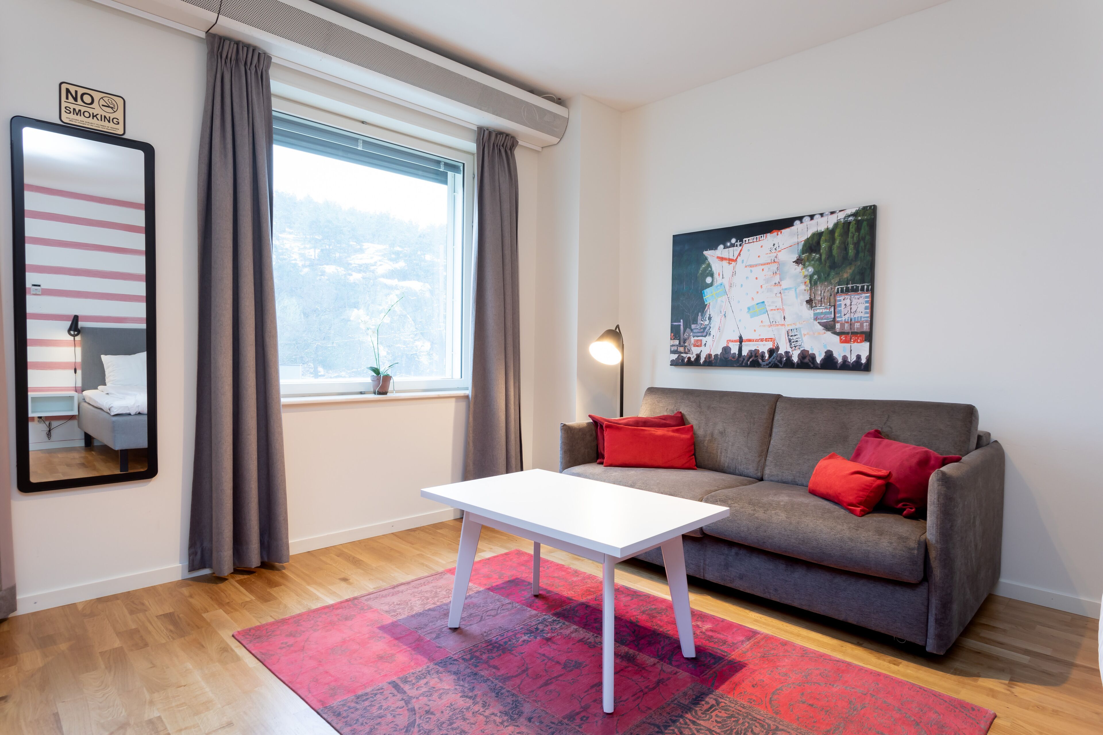 A modern living room with a grey sofa, red cushions, a white coffee table, a large window with curtains, a painting on the wall, and a "NO SMOKING" sign.