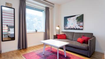 Ett modernt vardagsrum med en grå soffa, röda kuddar, ett vitt soffbord, ett stort fönster med gardiner, en tavla på väggen och en skylt med texten ”RÖKNING FÖRBJUDEN”.