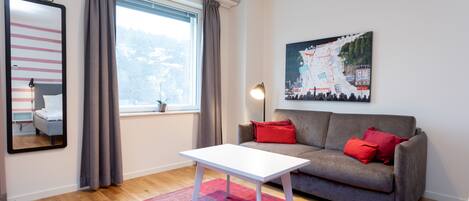 A modern living room with a grey sofa, red cushions, a white coffee table, a large window with curtains, a painting on the wall, and a "NO SMOKING" sign.