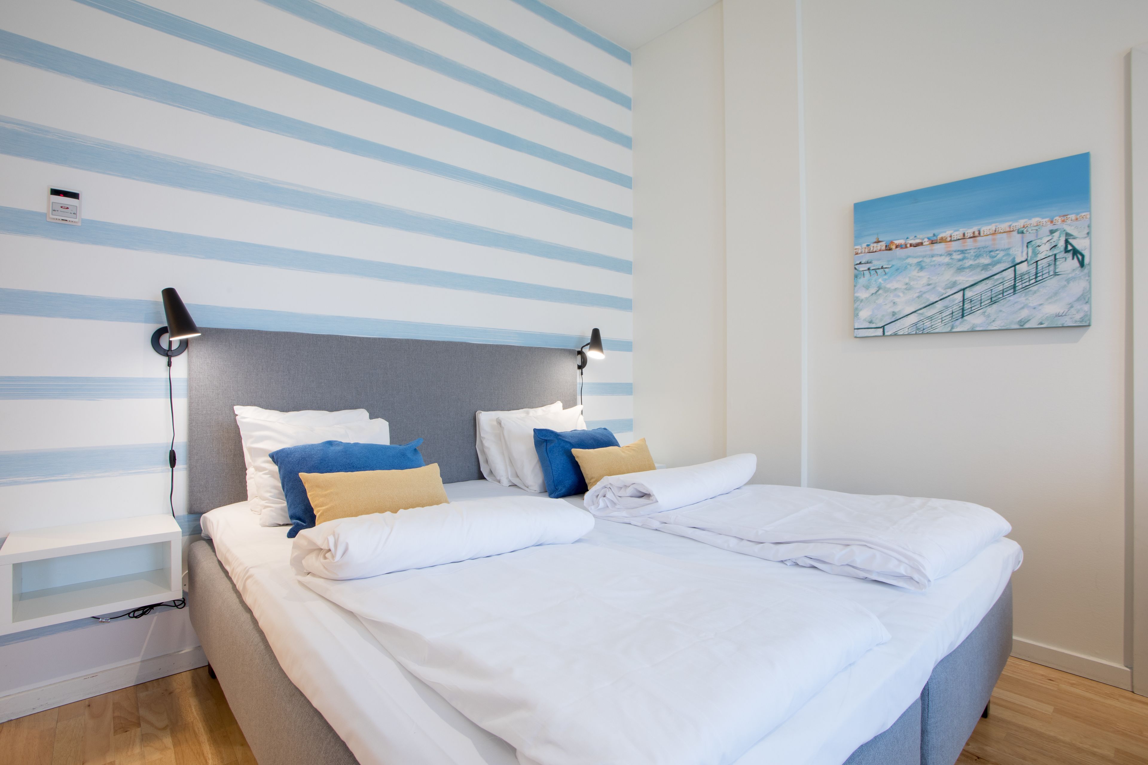 A neatly made bed with white bedding and blue and yellow pillows, a bedside table with a lamp, and a painting of a snowy landscape on the wall.