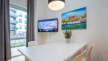 A modern hotel room with a large window, a flat-screen TV, a dining table with chairs, a potted plant, and a painting of a cityscape.