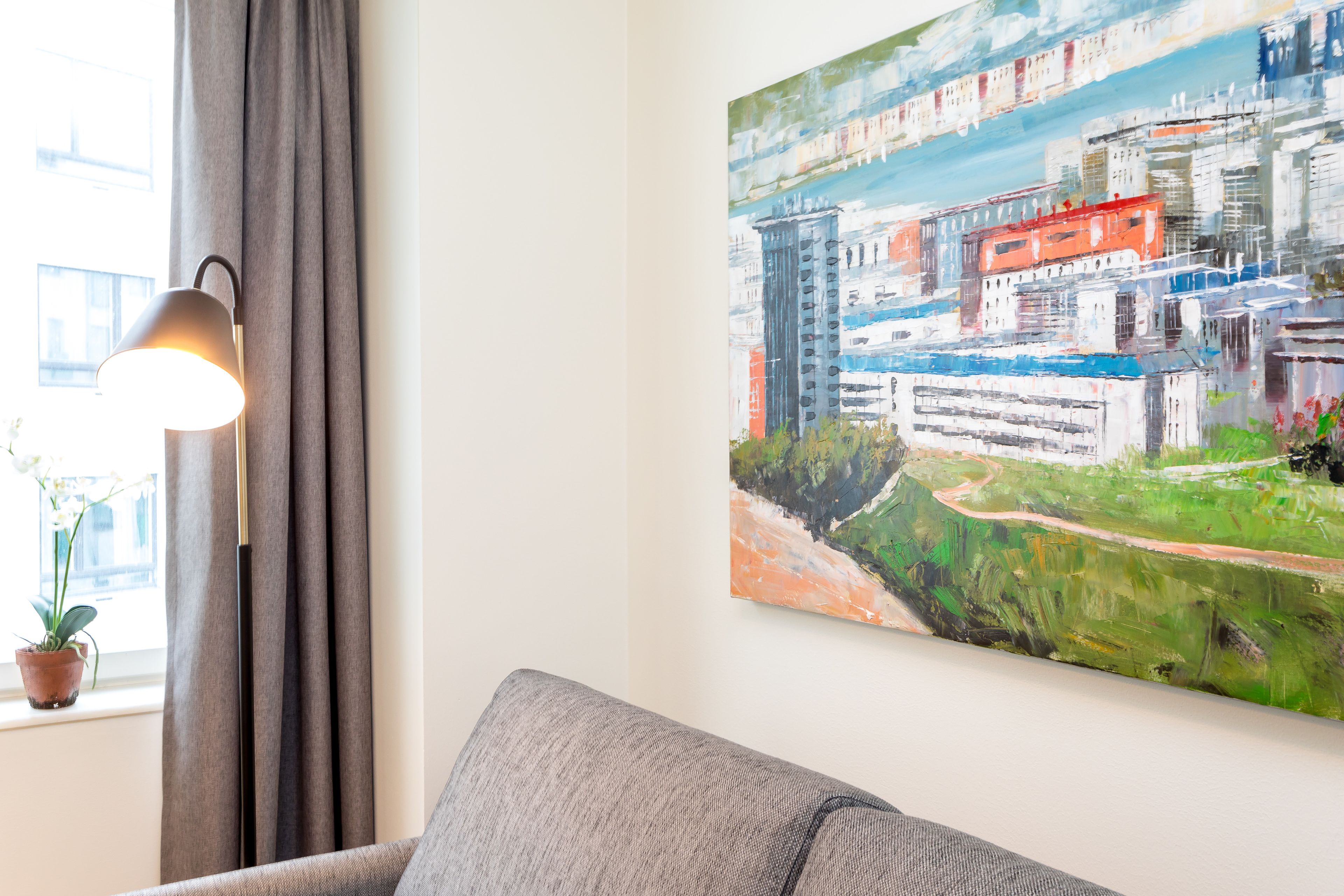 Una habitación con un sofá gris, una lámpara, un cuadro de un paisaje urbano y una ventana con cortinas.