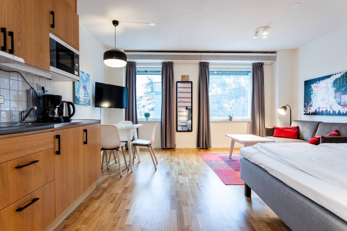 A modern hotel room with a wooden kitchenette, a dining area, a large bed, and a view of the outdoors.