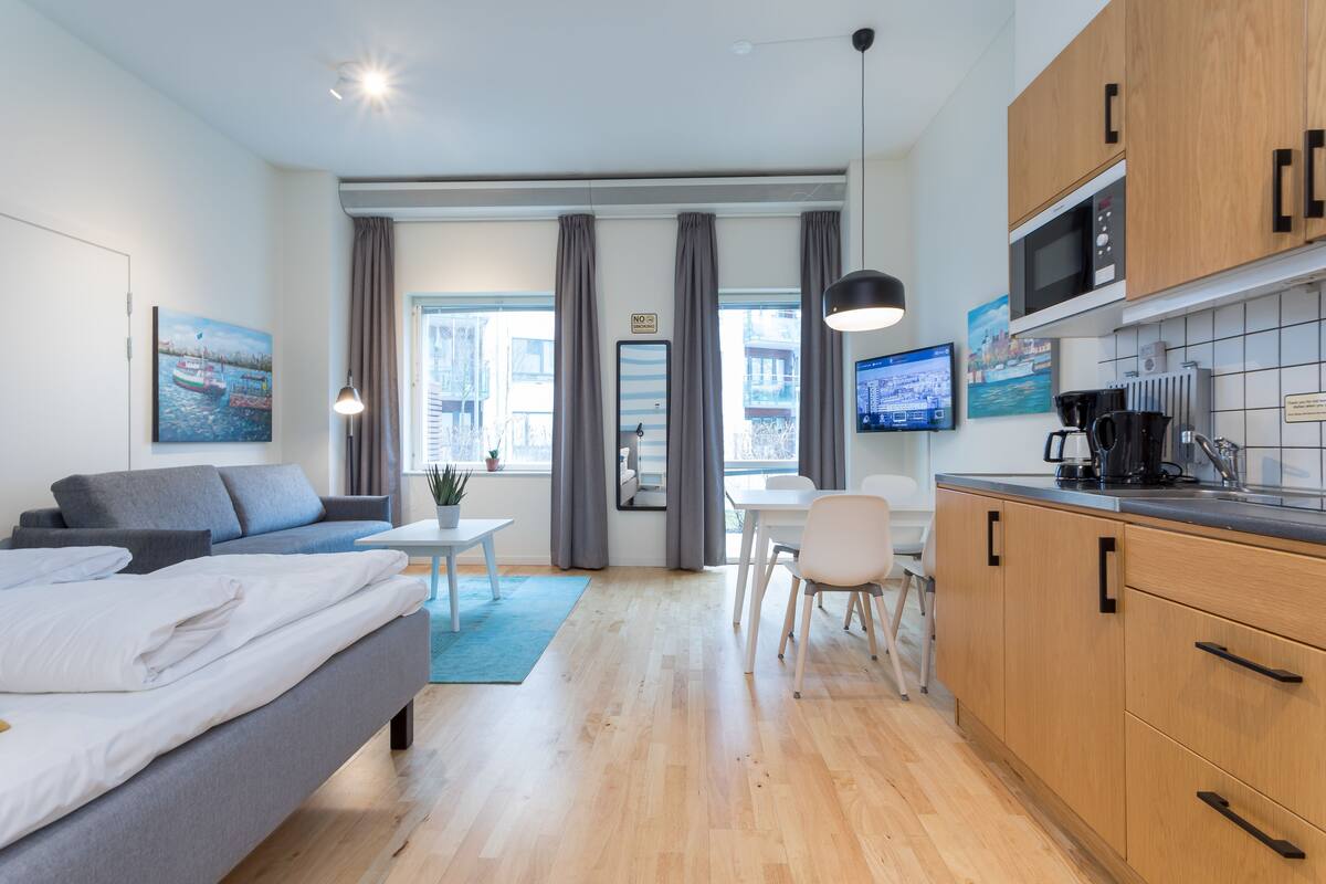 A modern hotel room with a bed, a sofa, a dining table, and a kitchenette.
