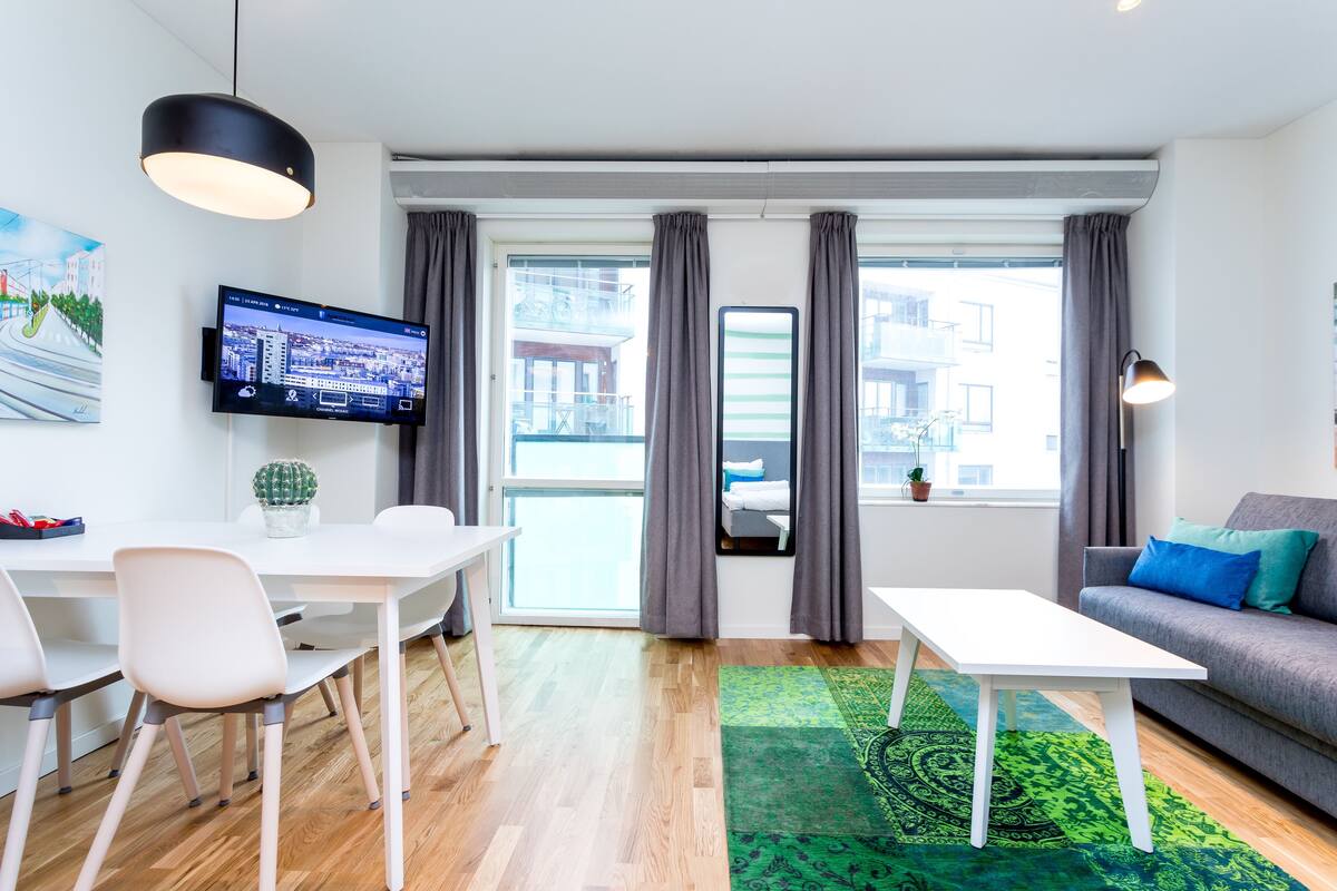 A modern living room with a flat-screen TV, a dining table with chairs, a sofa, a coffee table, and a green patterned rug.