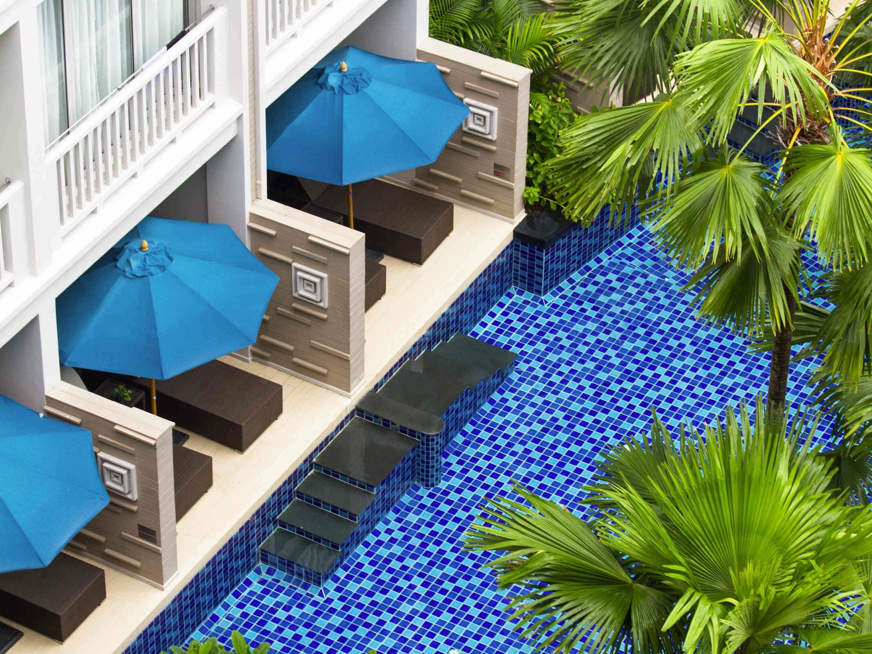 Un espace aménagé au bord de la piscine, avec des parasols bleus, une piscine carrelée et des palmiers.