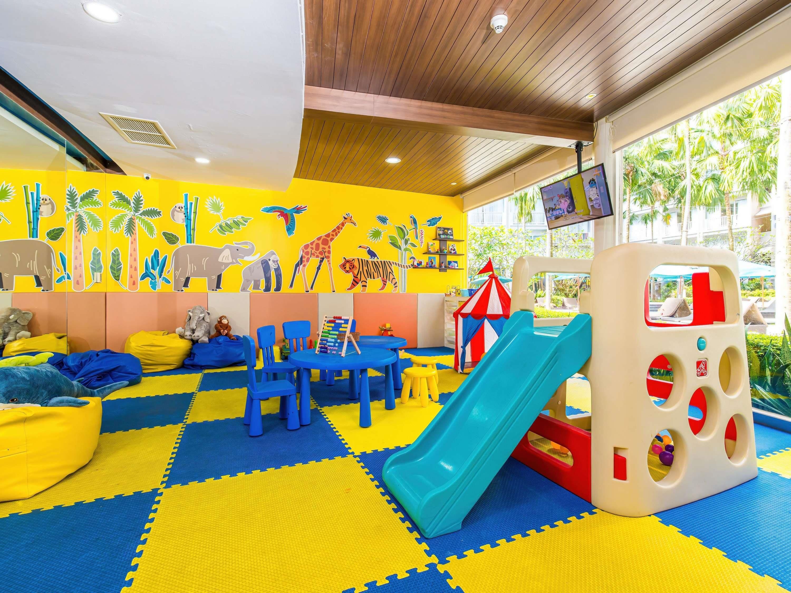 Zona de juegos infantiles con un tobogán, asientos y una pared decorada con ilustraciones de animales.