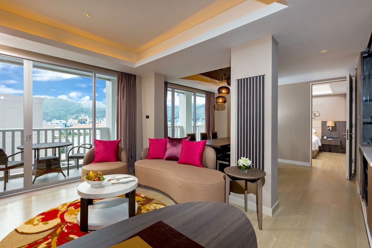A modern hotel room with a sofa, dining table, and a balcony with a city view.