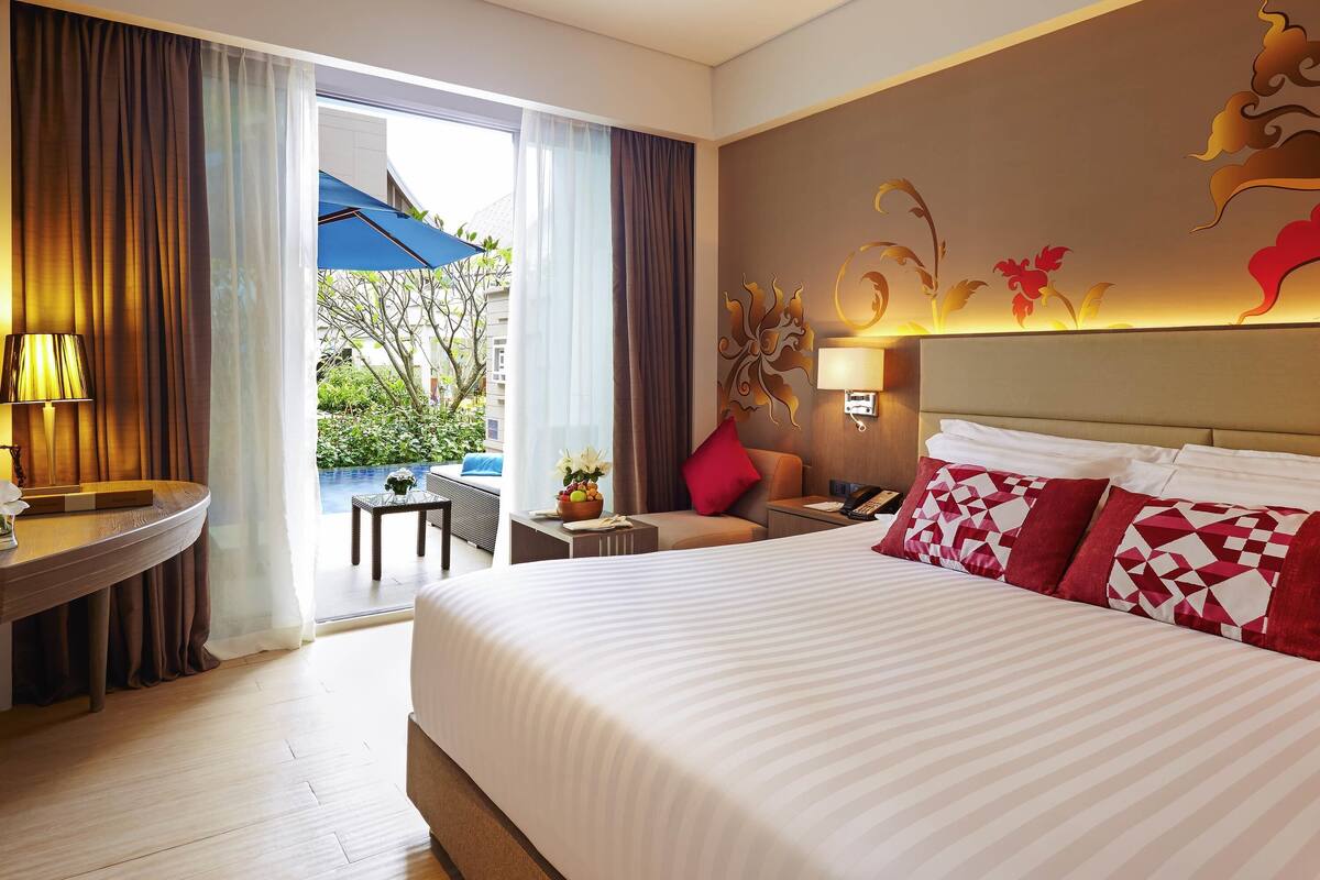 Une chambre d’hôtel avec un grand lit, un bureau avec une lampe, une petite table avec un vase, et une vue sur une piscine et la verdure environnante.