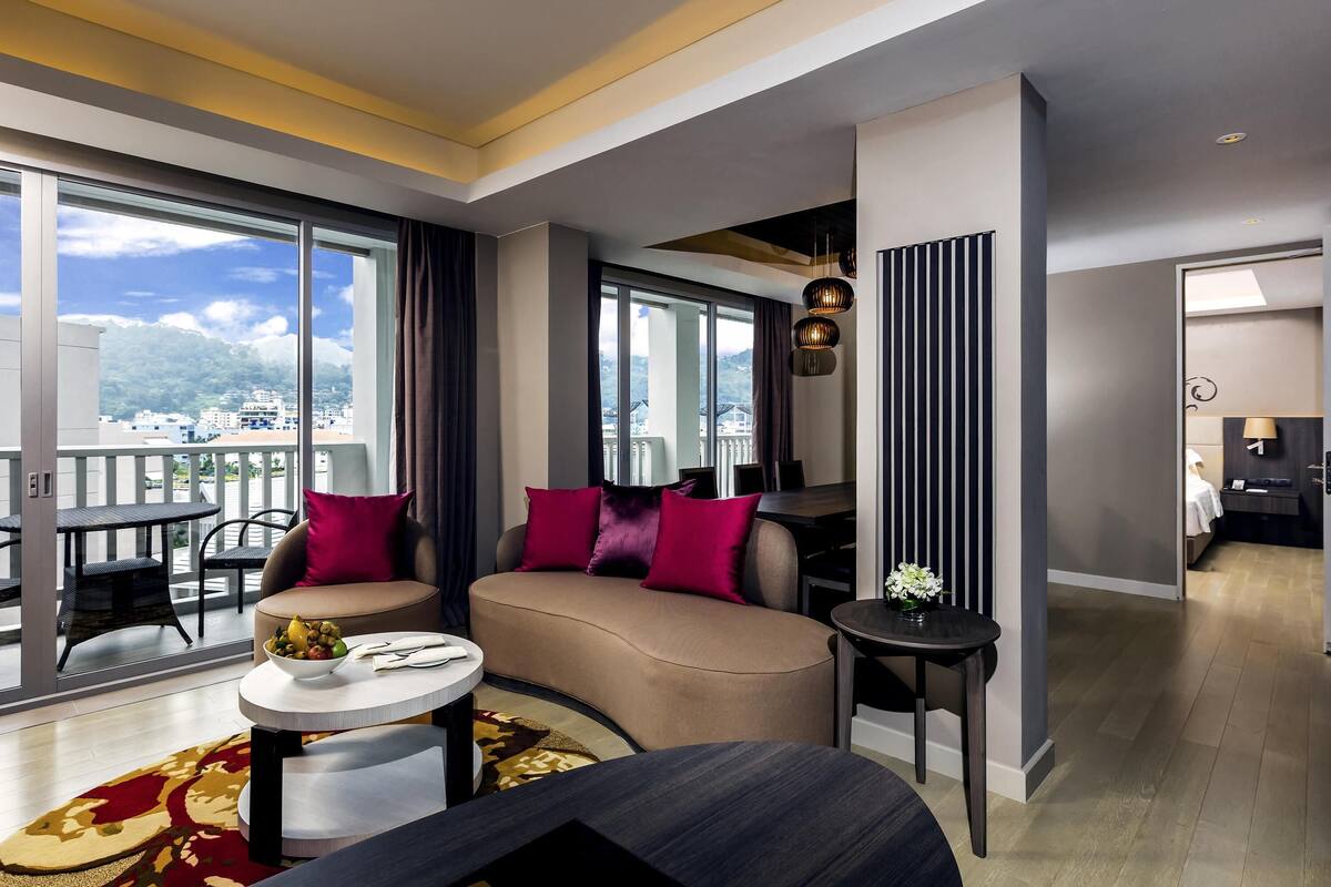 Une chambre d’hôtel moderne dotée d’un grand balcon, d’un canapé avec des coussins rouges, d’une table à manger et d’une petite table ronde agrémentée d’un vase de fleurs.