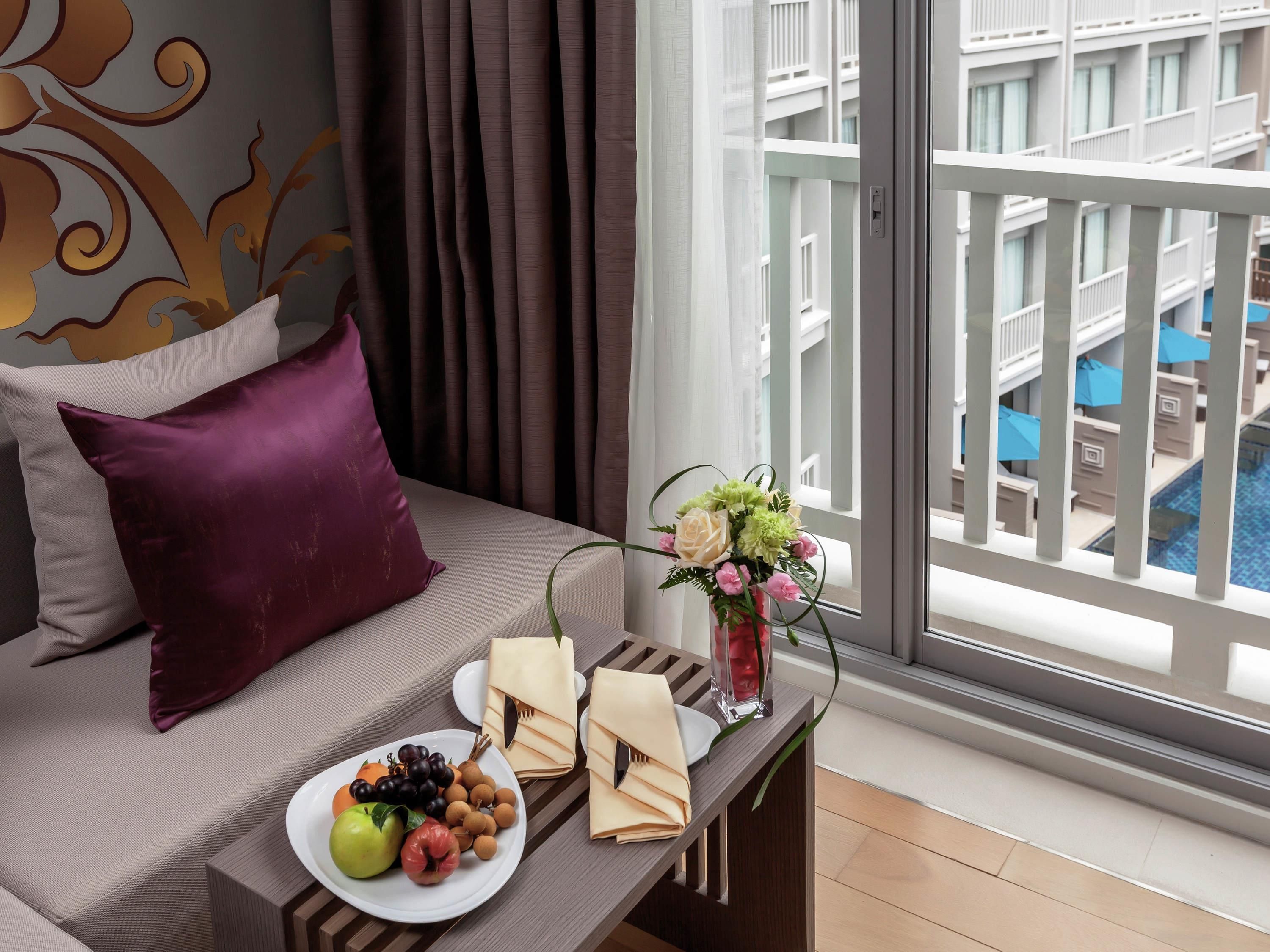Habitación de hotel con cama, una mesa de comedor con una bandeja de frutas, un jarrón con flores y vistas a una piscina y edificios.