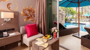 A hotel room with a sofa, a small table with a fruit plate, a vase with flowers, and a view of an outdoor pool area.