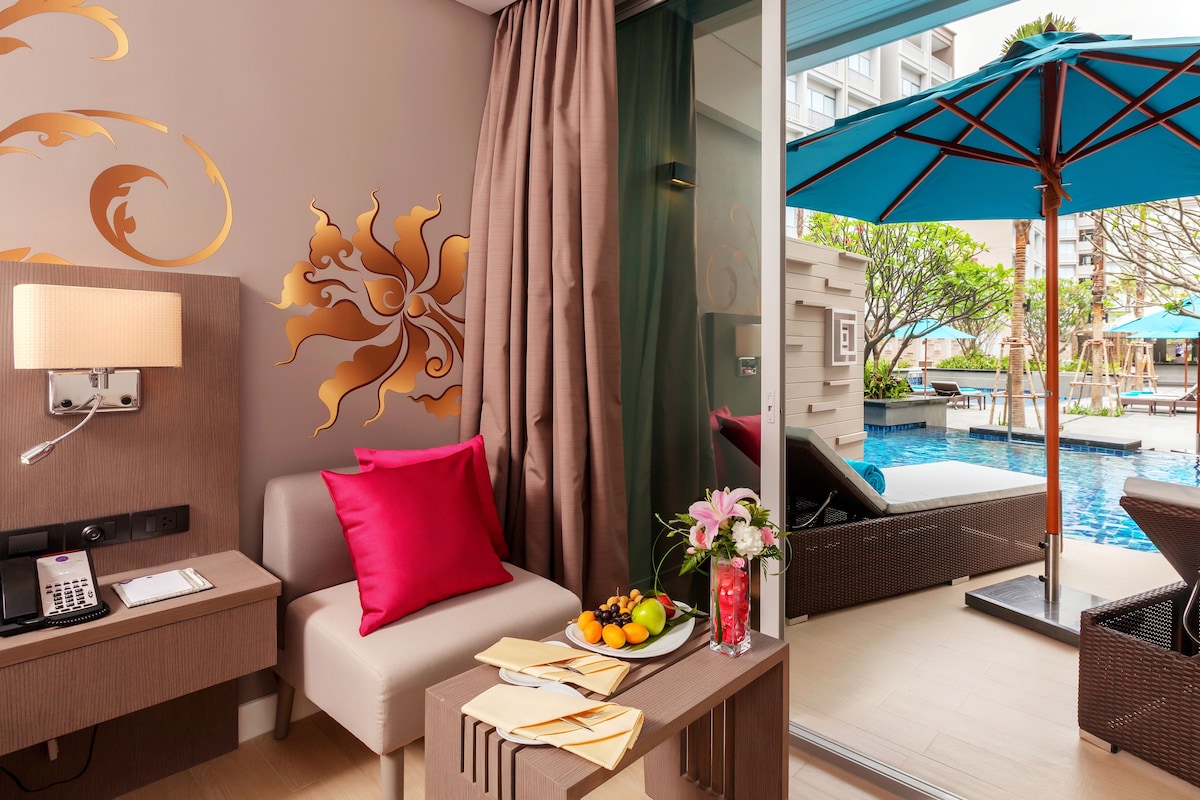 A hotel room with a sofa, a small table with a fruit plate, a vase with flowers, and a view of an outdoor pool area.