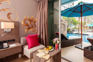 Une chambre d’hôtel avec un canapé, une petite table sur laquelle est disposée une assiette de fruits, un vase avec des fleurs et une vue sur l’espace extérieur abritant une piscine.