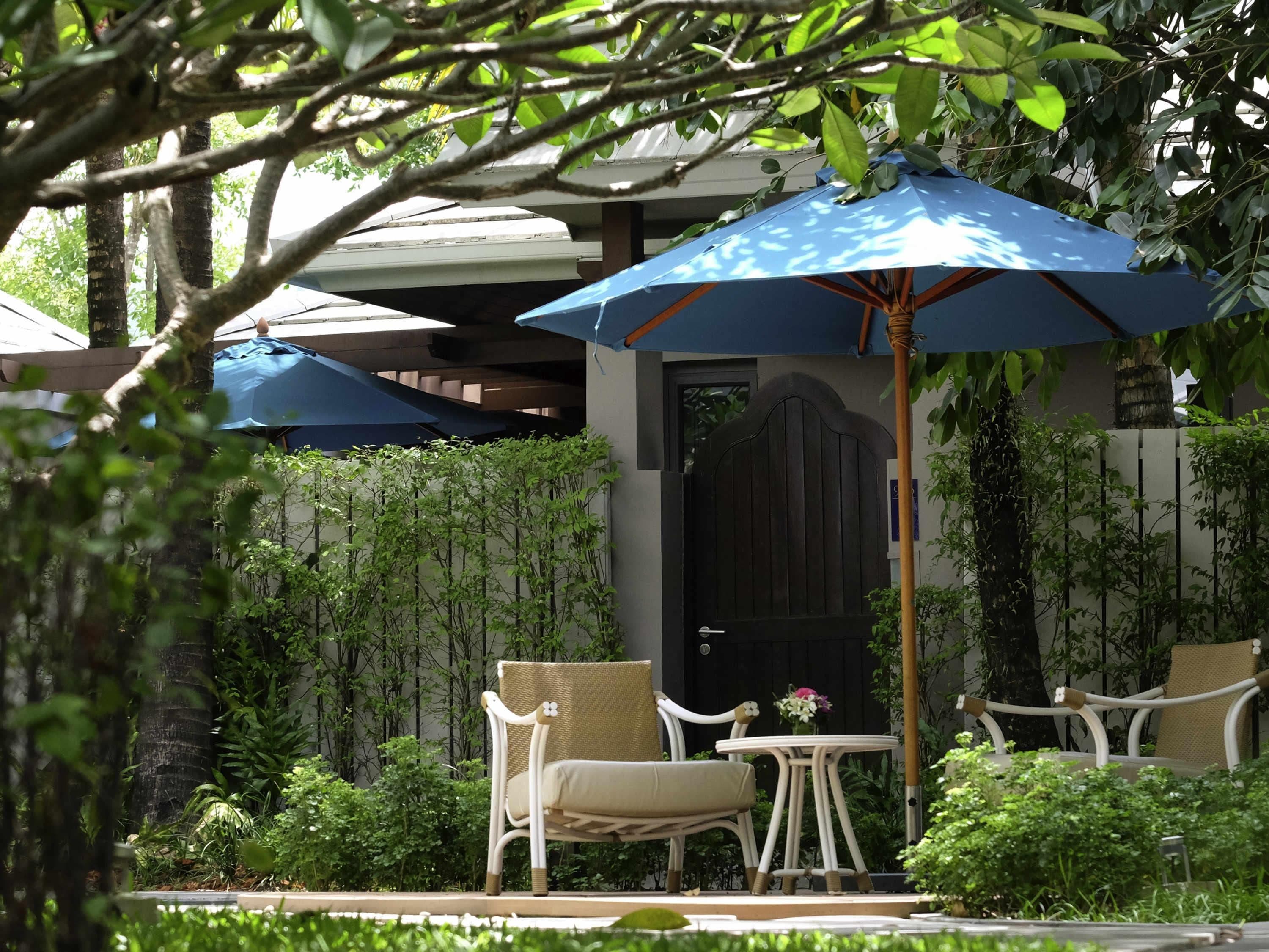 An outdoor seating area with a table and chairs under a blue umbrella, surrounded by greenery and a wooden fence.