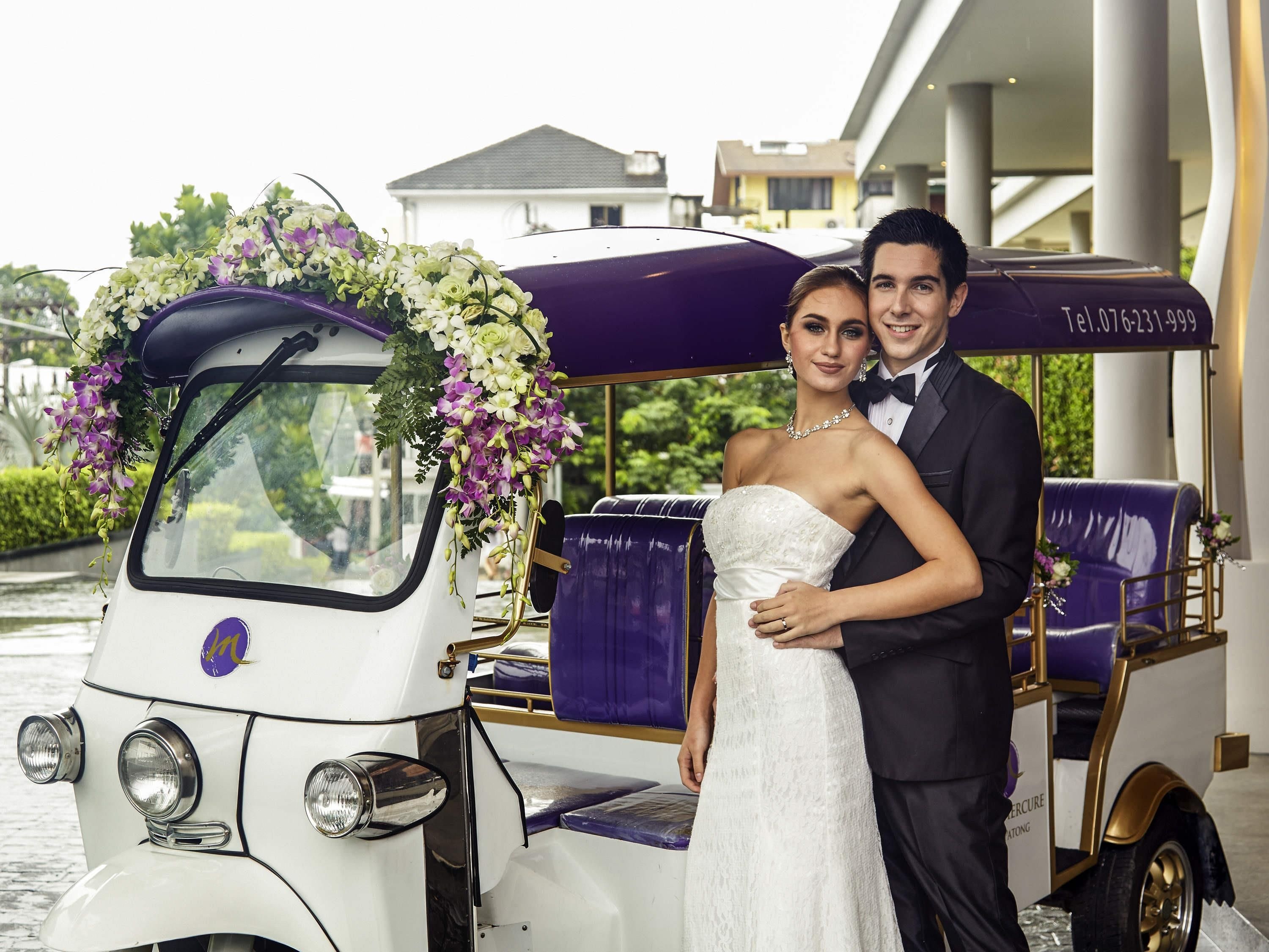 A wedding couple in a white wedding dress and black suit, standing next to a vintage car decorated with flowers.