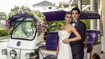 A wedding couple in a white wedding dress and black suit, standing next to a vintage car decorated with flowers.