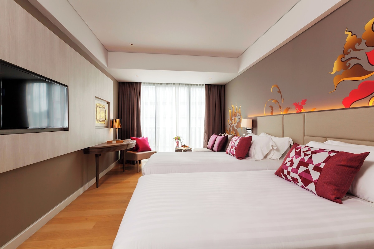 A hotel room with two beds, a TV, a desk, and a large window.